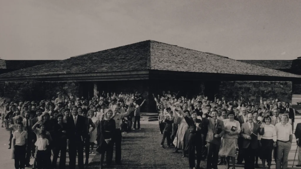 Black-and-white photograph of a large group of KCM Staff gathered outdoors in front the Kenneth Copeland Ministries Chapel building, many with arms raised and waving toward the camera.