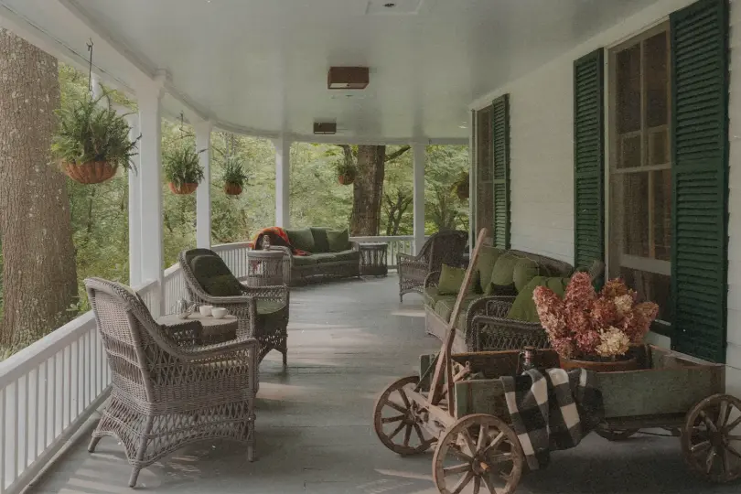 A covered front porch with wicker chairs and a matching sofa arranged along painted wooden floorboards, green cushions on the seating, and hanging plants suspended from the ceiling. A small wooden wagon filled with flowers and a folded blanket sits near a window with green shutters, while trees and greenery are visible beyond the porch railing.