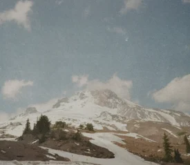 Snow-covered mountain peak under a bright blue sky with scattered clouds and evergreen trees at the base
