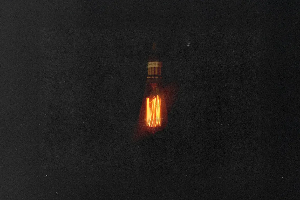 Single glowing Edison-style light bulb suspended in darkness, casting a warm orange filament glow