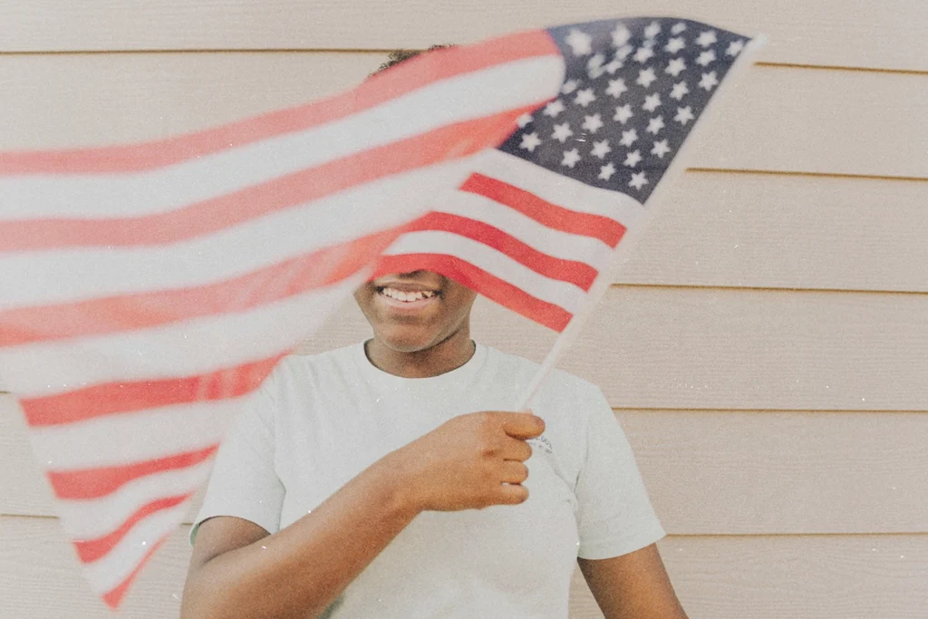 Young kid standing against a light-colored wall, waving an American flag that fills the foreground