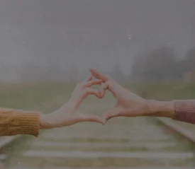 Two people extending their arms to form a heart shape with their hands over blurred railroad tracks