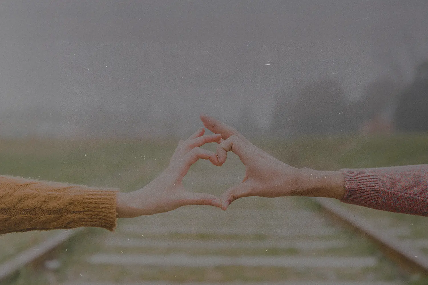 Two people extending their arms to form a heart shape with their hands over blurred railroad tracks