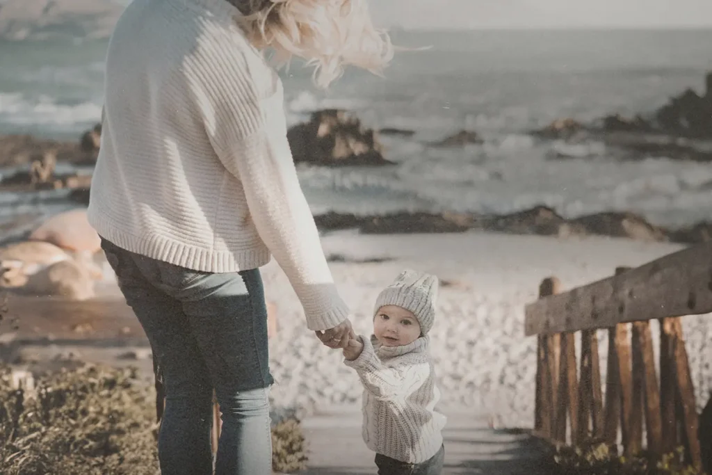 Adult holding a small child’s hand while walking down a wooden path toward a rocky beach with waves in the background