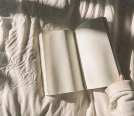 An Open Book is laid on a bed with a hand about to turn to the next page.