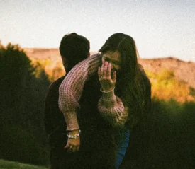Two people outdoors, one carrying the other on their back in a grassy area with trees and hills under a clear sky.