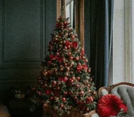 Decorated Christmas tree with red bows and ornaments beside a window and vintage sofa.