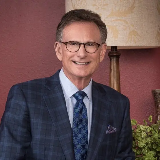 A man wearing a suit and glasses seated indoors against a warm-colored background.