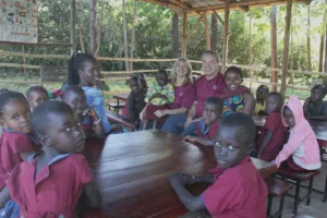 Children gather around missionaries seated at outdoor tables during a Uganda missions class, reflecting their commitment to teaching and discipleship.