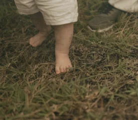 A small child’s bare feet standing on grass next to an adult’s shoes.