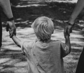 A child walking outdoors while holding the hands of two adults on each side.