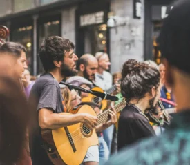 A street musician playing guitar during an outdoor performance, surrounded by people in a city area.