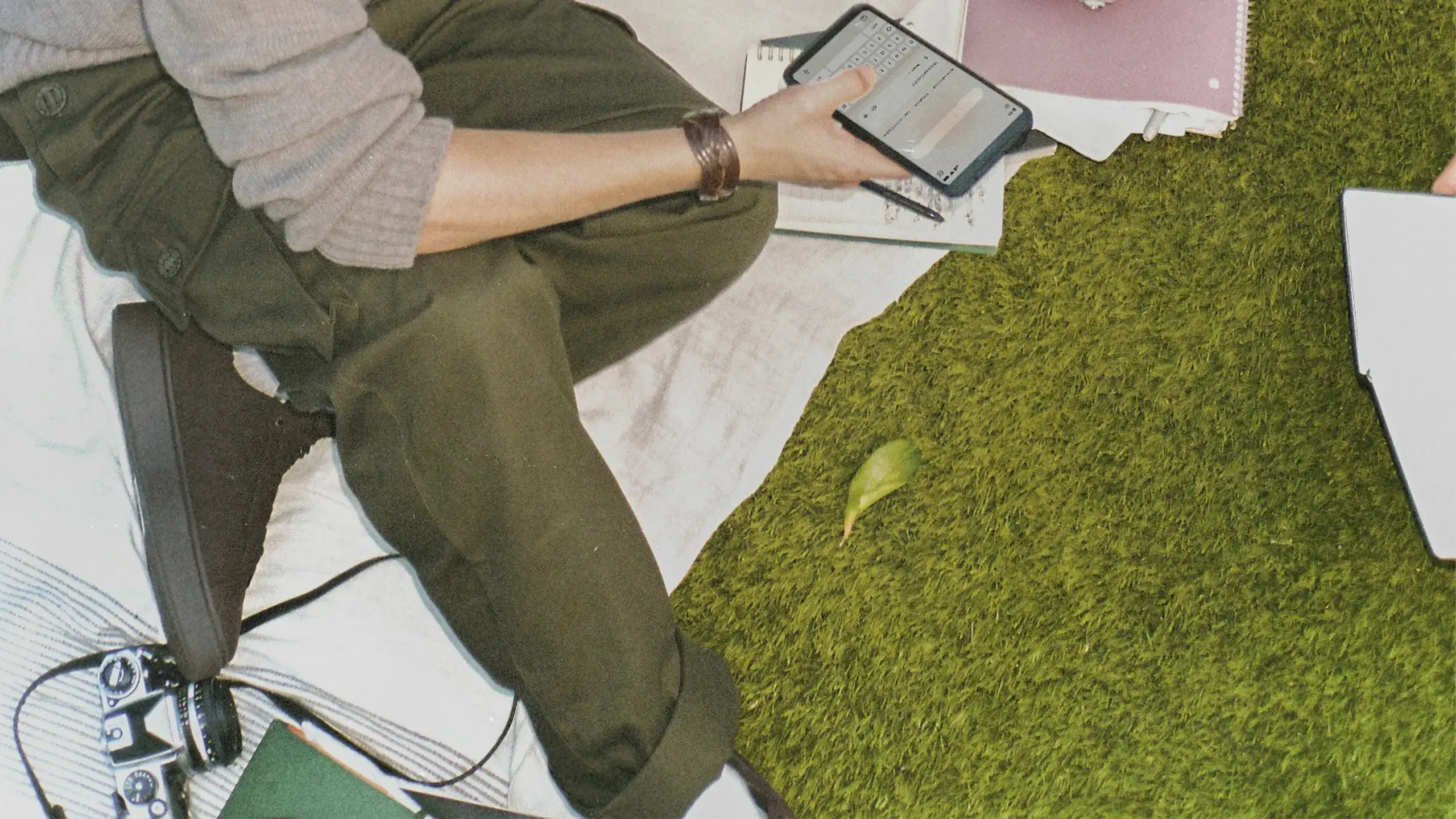 Person sitting on grass and paper notes, holding a smartphone with notebooks and a laptop nearby in an outdoor work or study setup